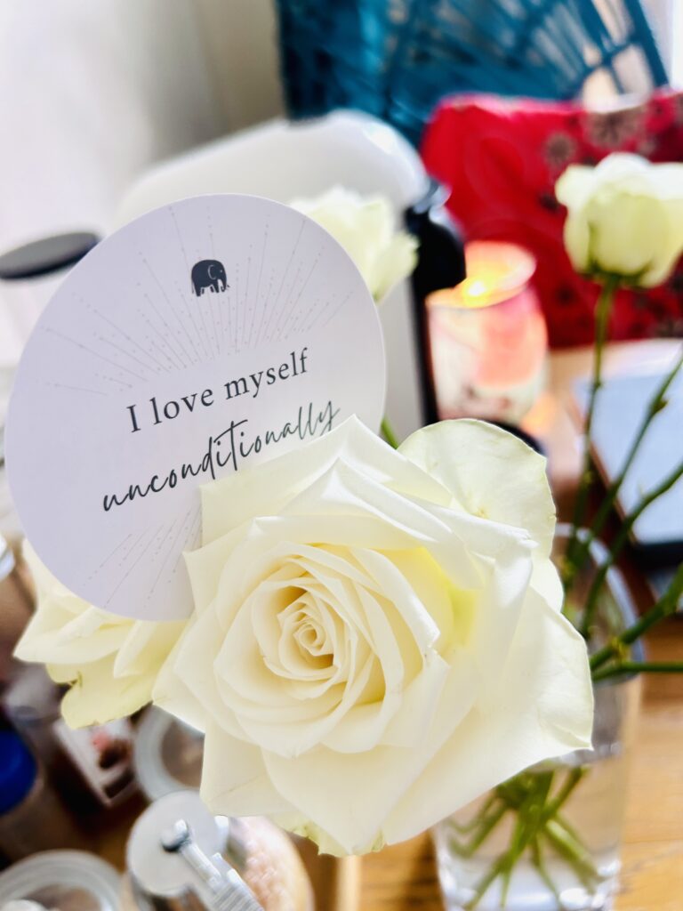 Image shows a picture of an affirmation card reading 'I love myself unconditionally'.  It is balanced on a cream rose.